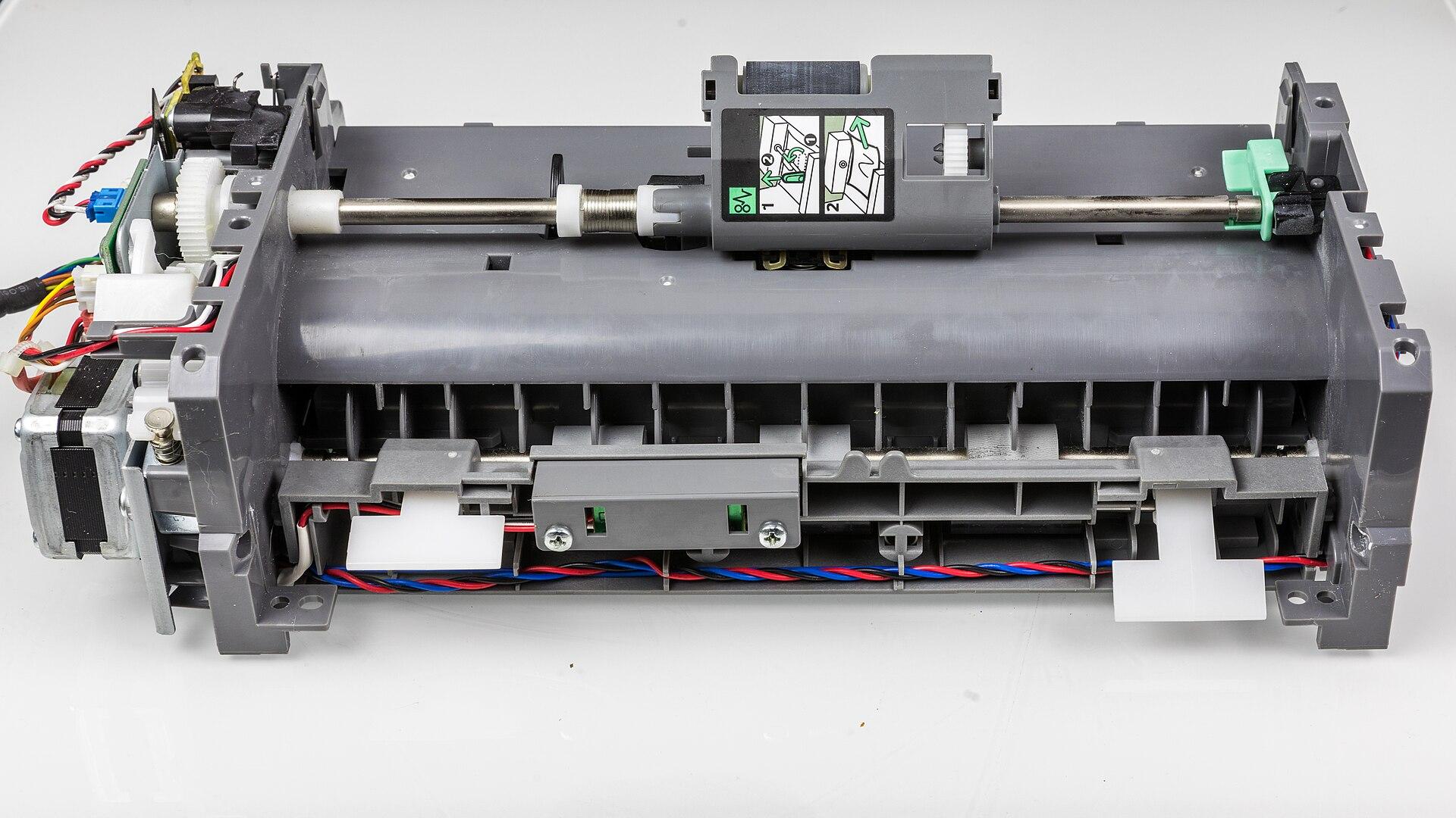 Automatic document feeder assembly removed from a machine and photographed on a neutral background, showing feed rollers on a drive shaft, a separation section, gear train, stepper motor and wiring harness