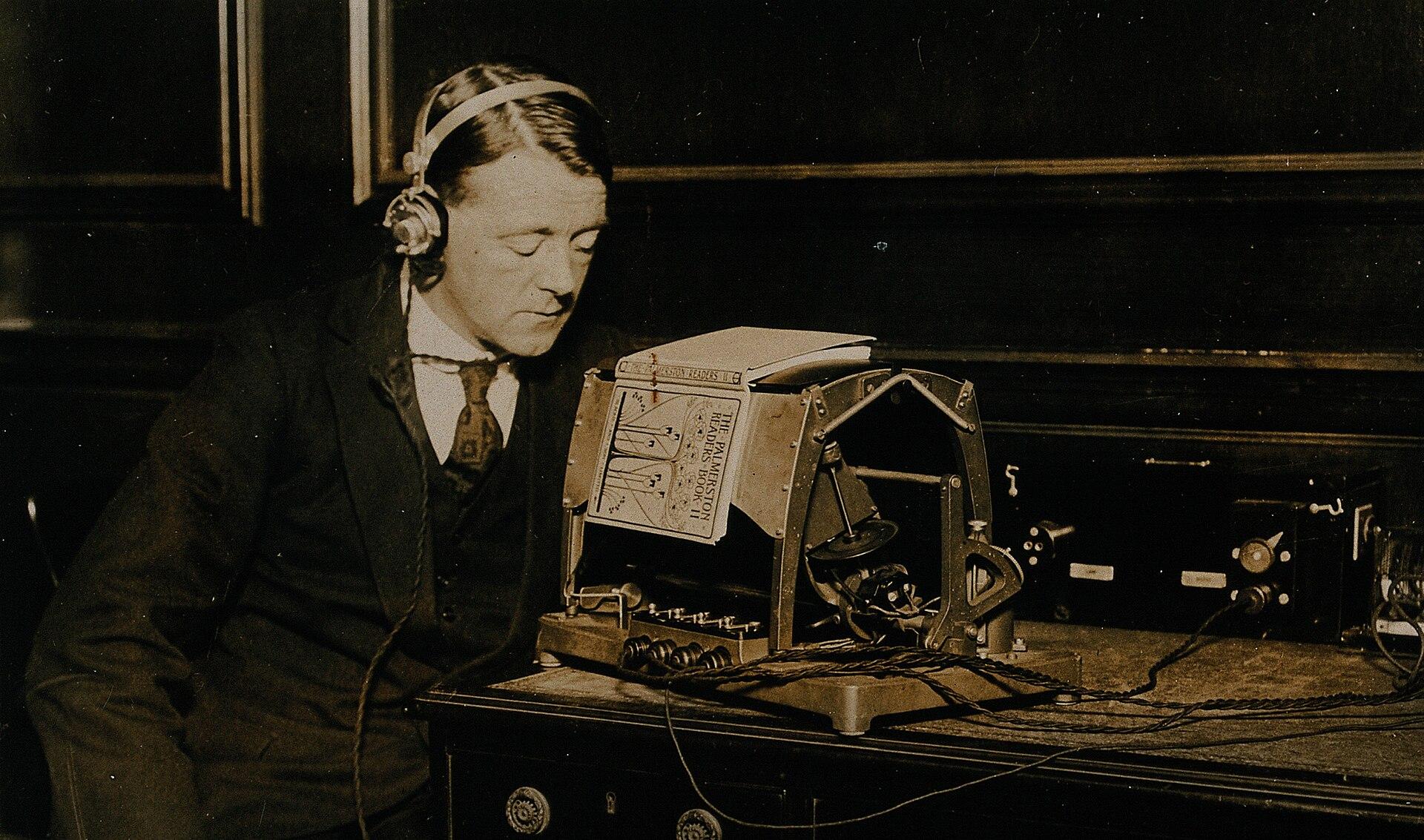 Photograph from about 1921 of a man demonstrating the optophone, a reading device for blind users, seated at an apparatus with an optical head over a page