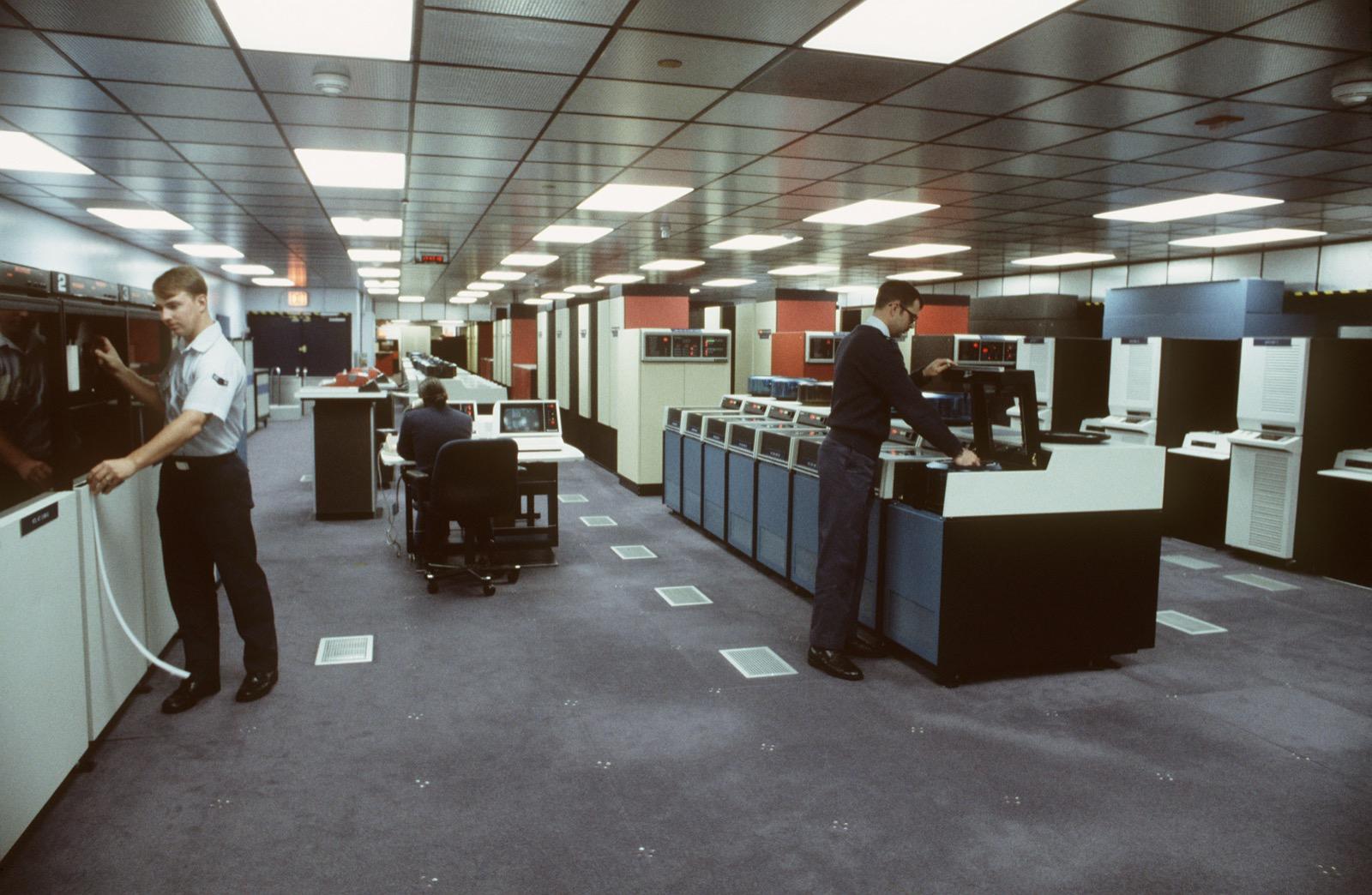 Operators seated at terminal workstations inside the fluorescent-lit NORAD Computer Center command room, equipment racks lining the background