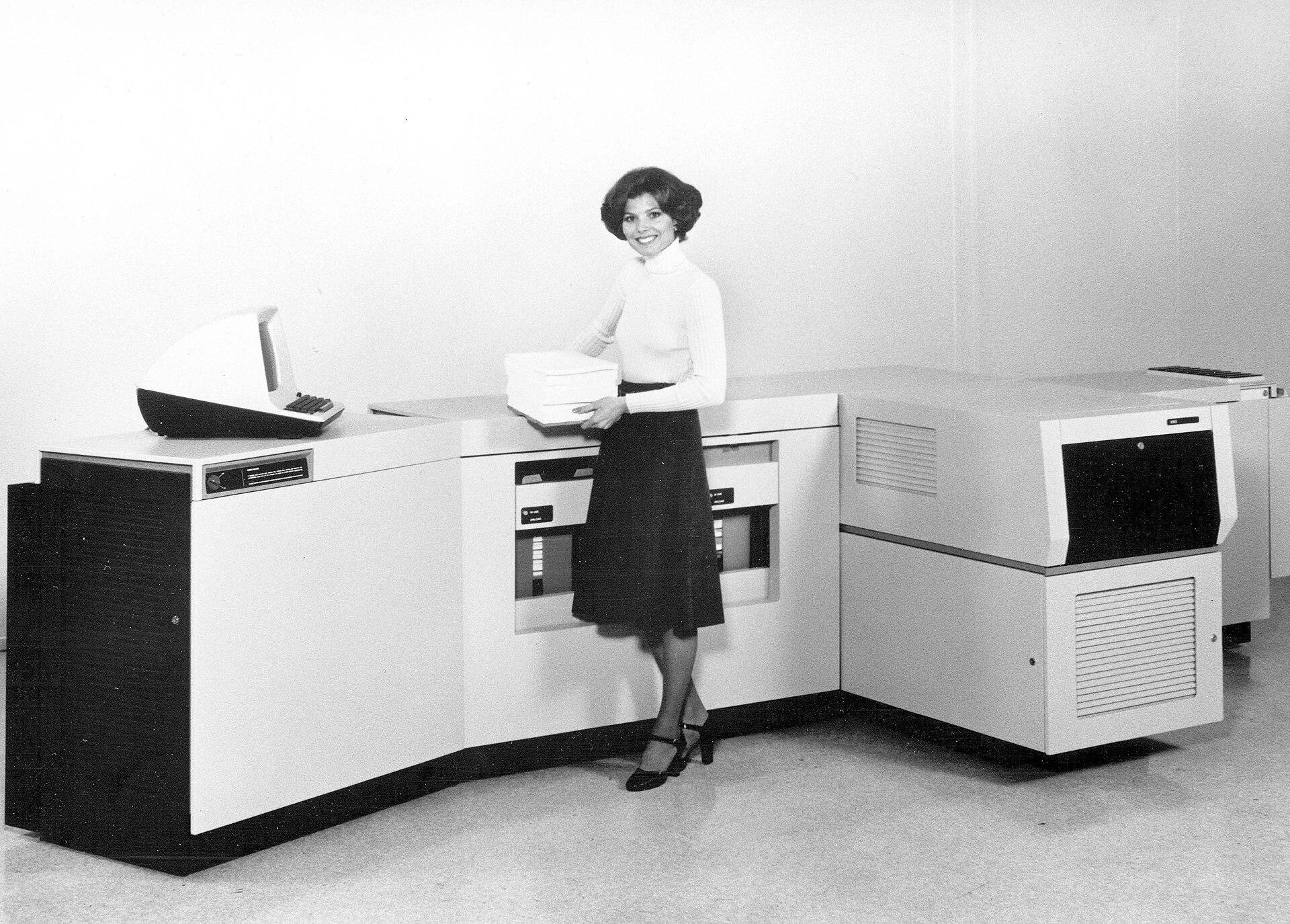1977 black-and-white publicity photograph of the Xerox 9700 Electronic Printing System, an L-shaped run of cabinets with an operator console at one end, a person standing beside it holding a stack of printed output