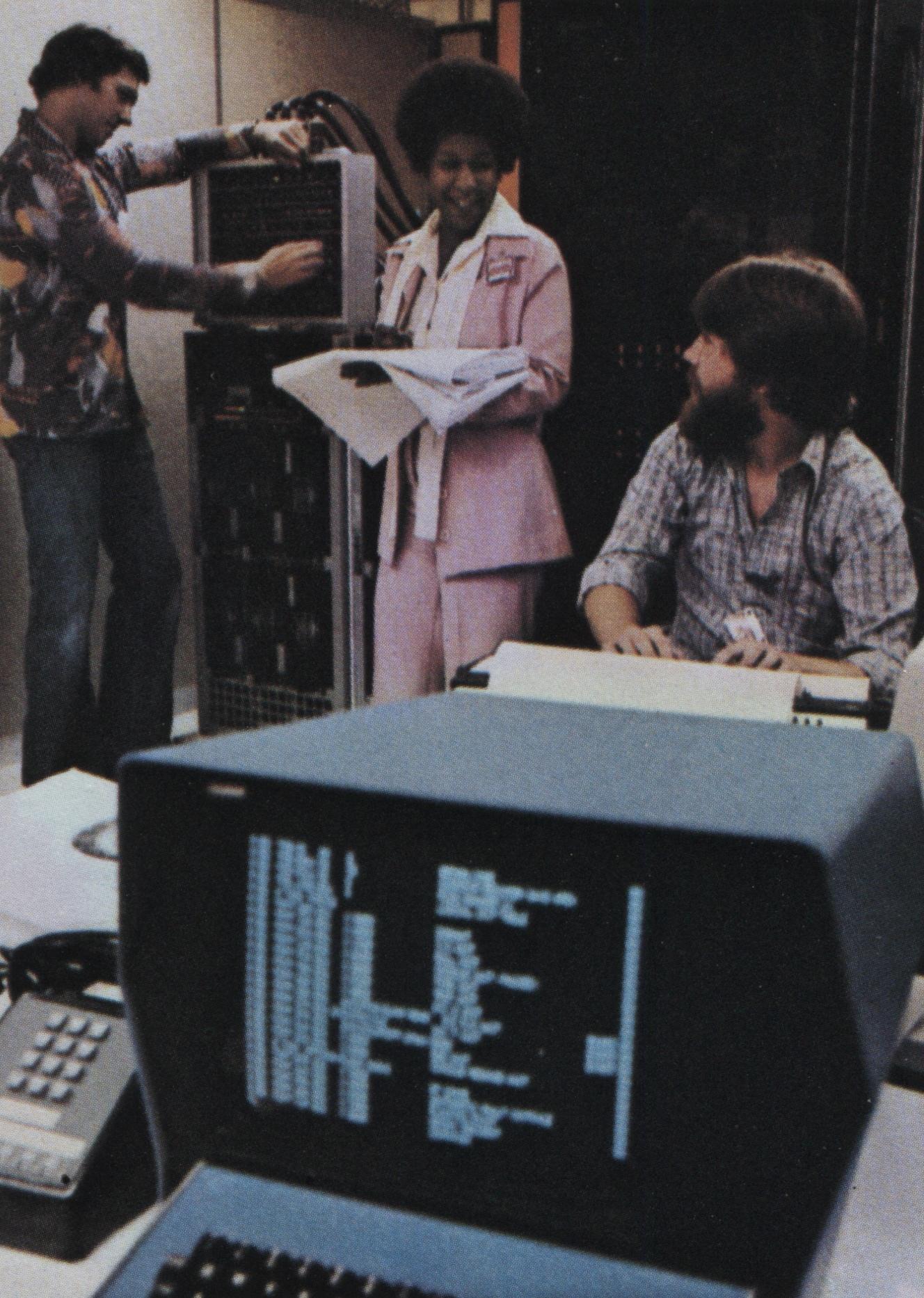 Three Hughes Aircraft employees in office attire in conversation directly in front of corporate mainframe equipment racks and tape units