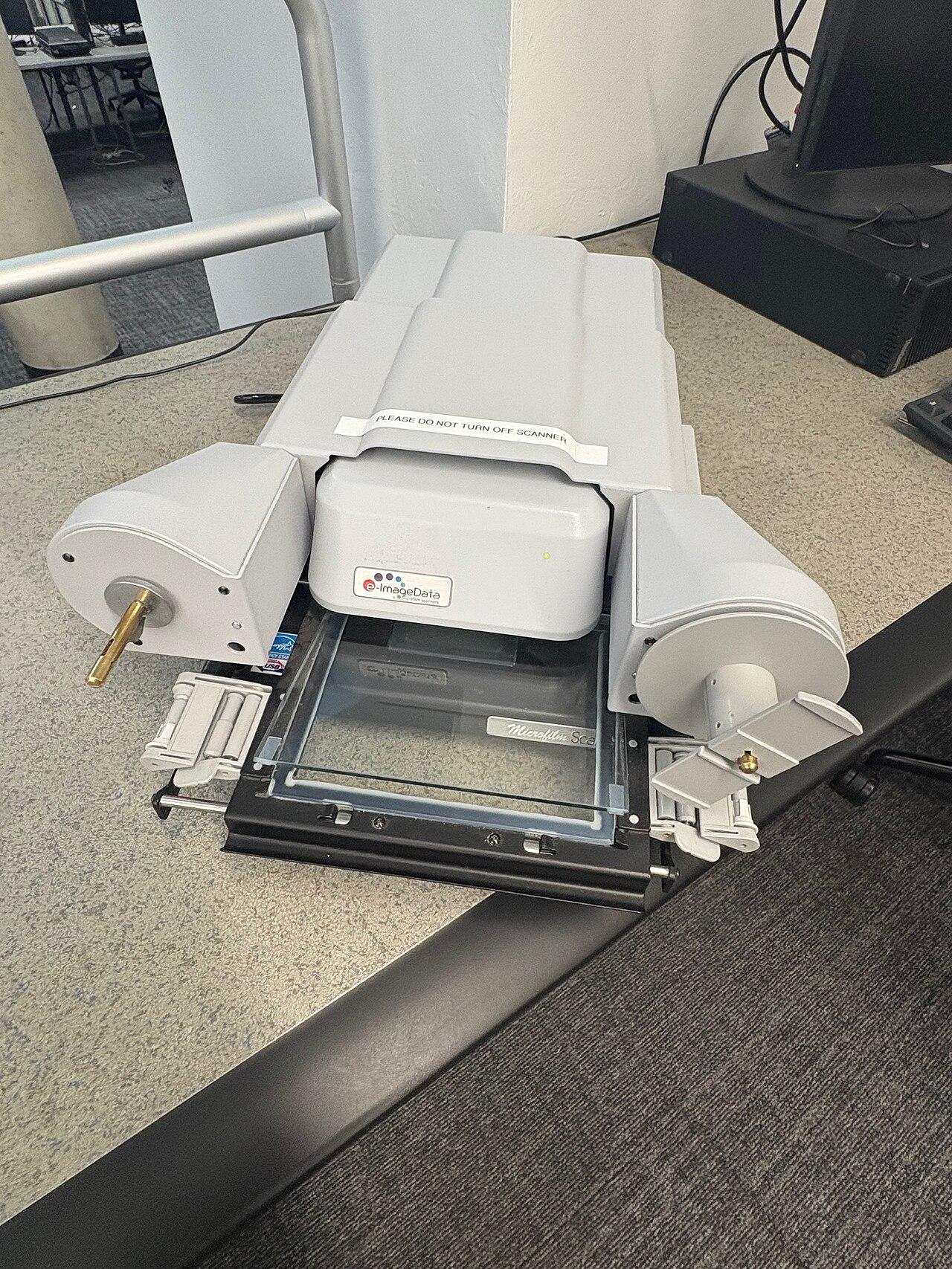 Microfilm reader-scanner: a desktop unit with a film transport beneath an optical head, connected to a display showing the scanned frame