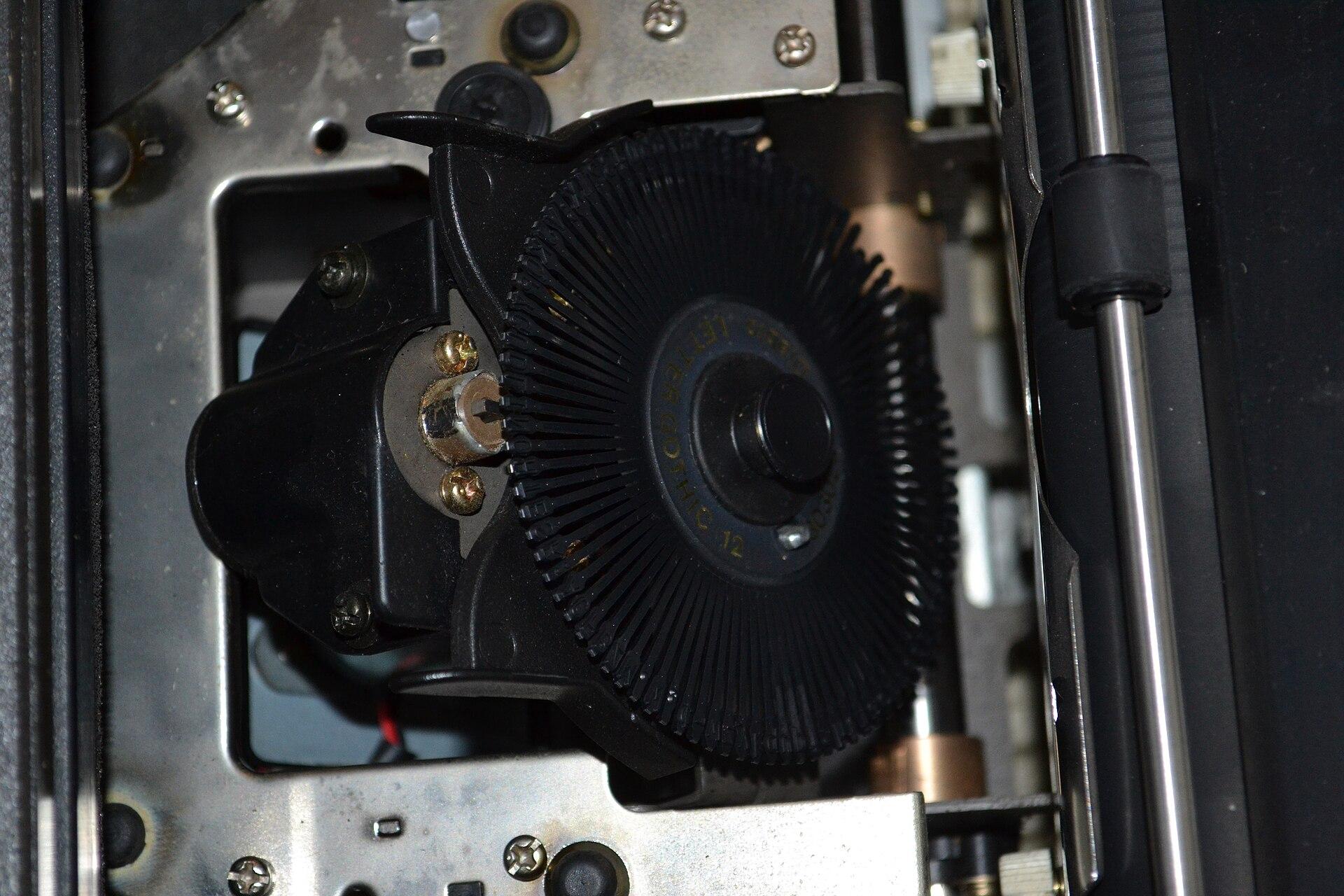 Close view of a daisy-wheel print head: a spoked wheel of type petals behind a ribbon cartridge, with the hammer positioned at the print point