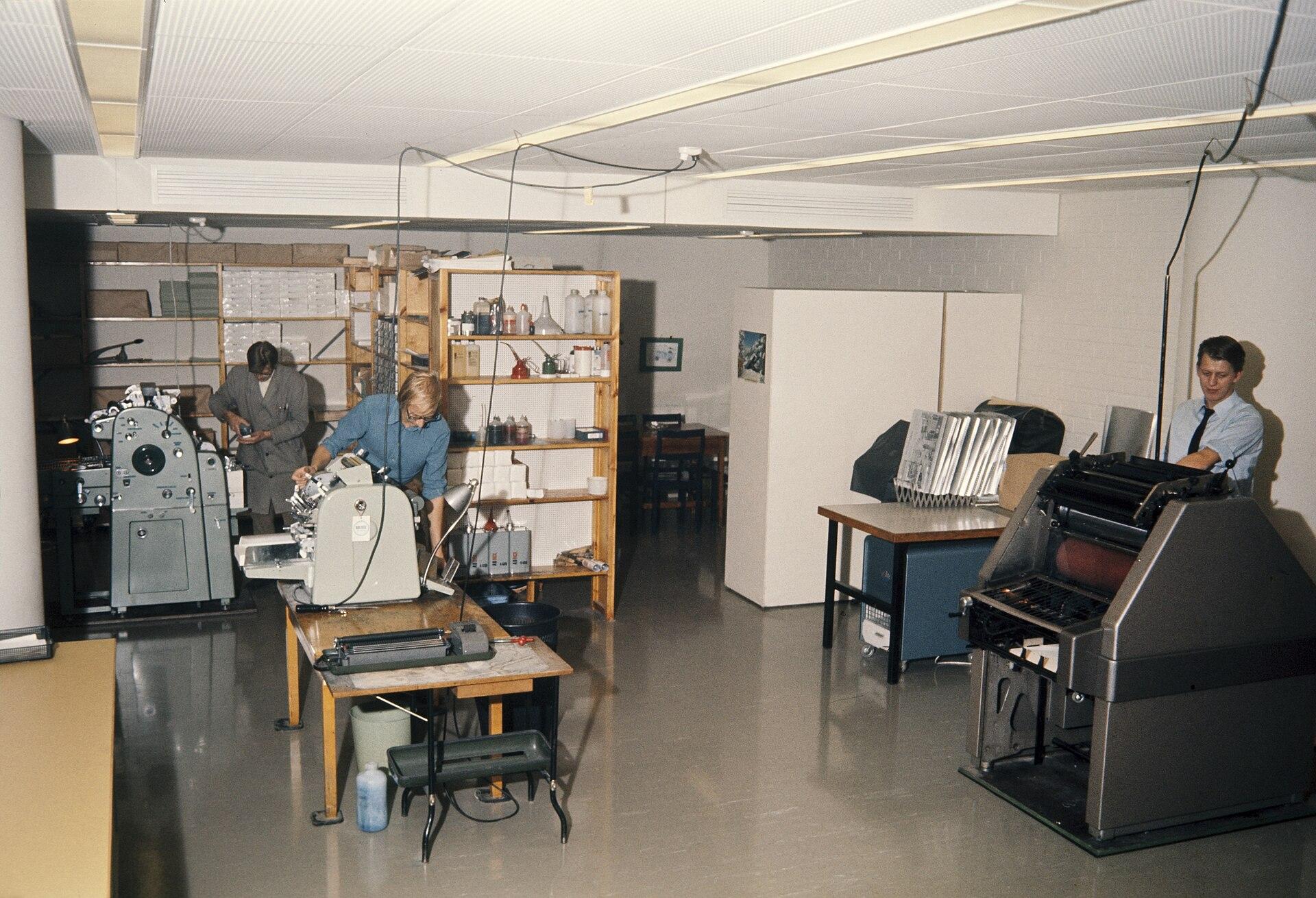 Colour photograph of a 1969 office duplicating room: a worker at a duplicating machine surrounded by paper stock, output trays and shelving
