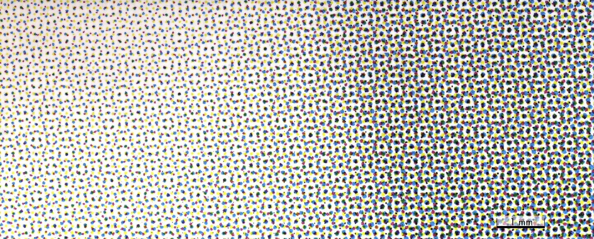 Magnified photograph of a printed CMYK halftone screen with a one millimetre scale bar, showing cyan, magenta, yellow and black dots forming rosettes and growing larger from light areas to dark
