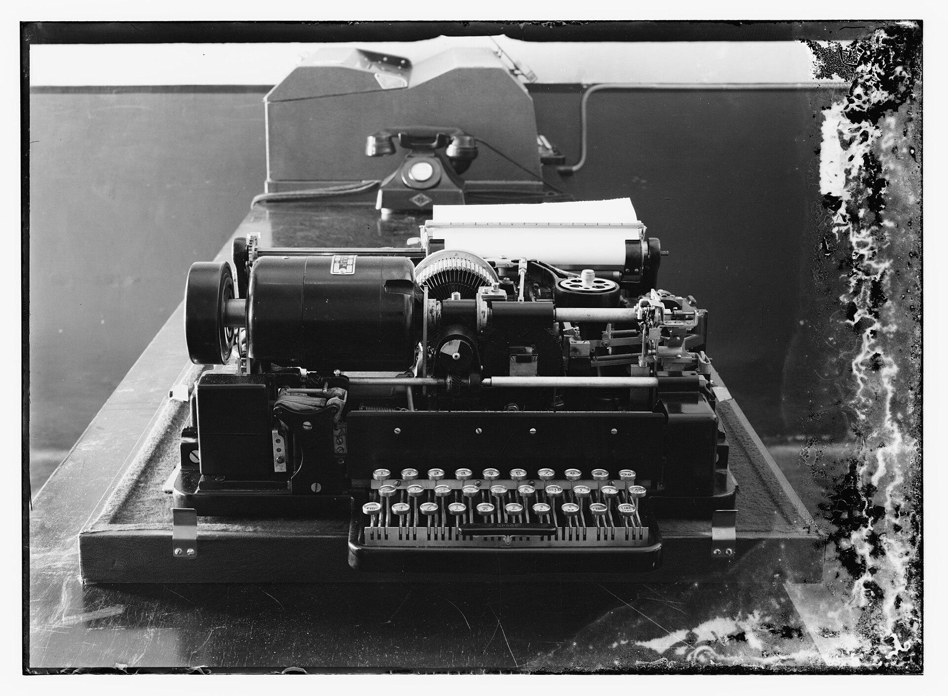 1938 photograph of a teleprinter with its mechanism exposed, showing keyboard, type mechanism, paper roll and drive motor, with a desk telephone behind it